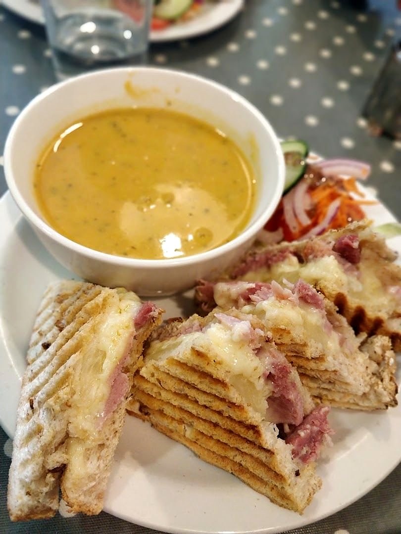 A bowl of soup with a ham-and-cheese toastie cut into pieces and a little salad, on a white plate on a spotted tablecloth